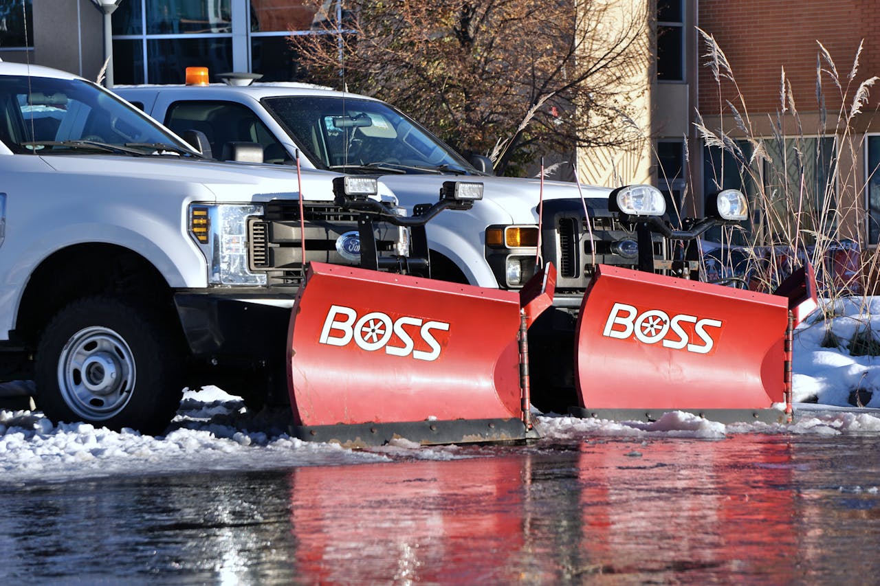 Two white pickup trucks equipped with red BOSS snow plows are parked on a snowy surface, reflecting in a wet patch. Some snow and bare trees are visible in the background.