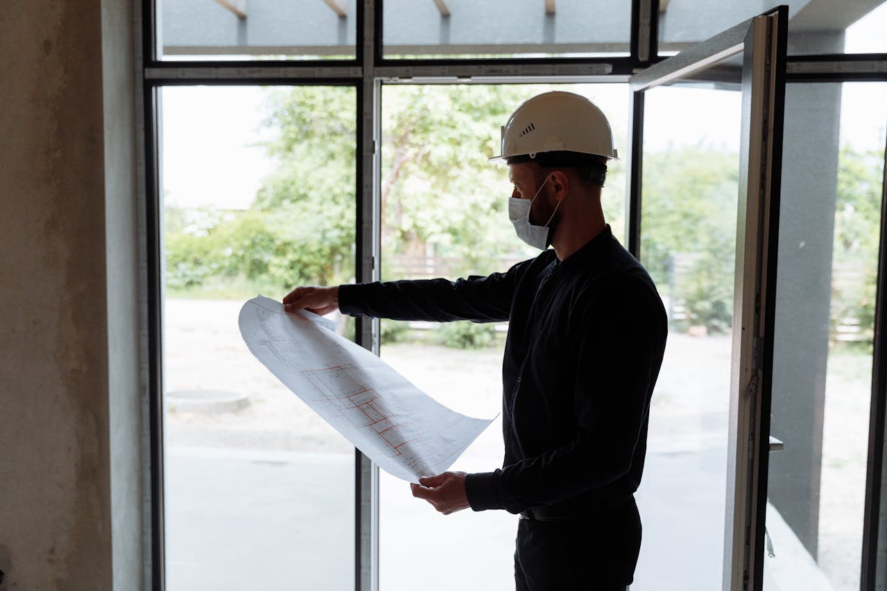 A person wearing a white hard hat and a face mask stands indoors by an open door, holding and examining a large sheet of architectural blueprints. Natural light comes in through the glass windows behind them.
