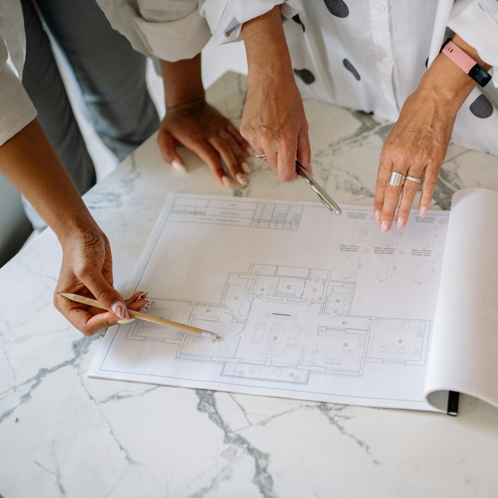 Two people discuss a building floor plan, pointing at the blueprint with a pen and pencil on a marble table. Only their hands and part of their arms are visible.