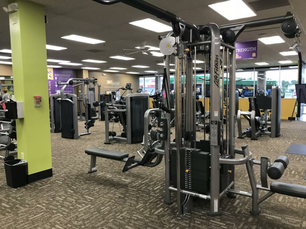 A spacious gym with various exercise machines, including weight machines and cardio equipment, on a brown carpeted floor. The area is brightly lit with green and purple accents on the walls.
