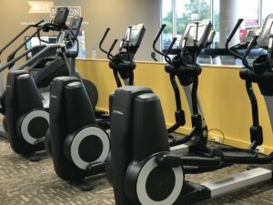 A row of elliptical machines and exercise equipment in a well-lit gym with carpeted floors, yellow walls, and large windows showing outdoor greenery.