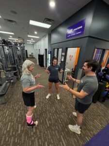 Three people stand in a circle inside a gym, talking and gesturing with their hands. Gym equipment and a purple sign reading "Coaching" are visible in the background. All three people are wearing athletic clothes.