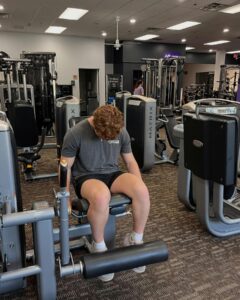 A person with short brown hair in a gray shirt and shorts sits on a leg extension machine in a gym, looking down. Exercise equipment and other gym-goers are visible in the background.