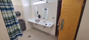 A public restroom with two sinks, large mirrors, a paper towel dispenser, a trash can, and a striped shower curtain. A door is partially open with a person barely visible behind it. Toiletries sit on the counter.