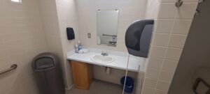 A small public restroom with a white sink, mirror, soap dispenser, paper towel dispenser, trash can, and a blue bucket beneath the sink, surrounded by beige tiled walls.