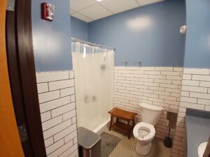 A clean bathroom with blue walls, white subway tile, a shower with a clear curtain, a toilet, a wooden bench, a trash can, a toilet paper holder, and a gray floor mat.