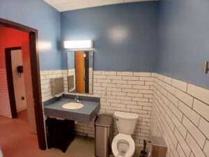 A public restroom with blue walls, white subway tile halfway up, a white toilet, a sink with a mirror above it, a trash can, and a partially open door leading to a hallway with orange lighting.