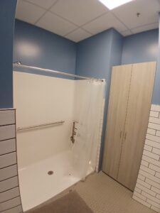 A bathroom with blue walls, a walk-in shower featuring a grab bar and folding seat, a white shower curtain, tiled lower walls, hexagonal floor tiles, and a tall wooden cabinet.