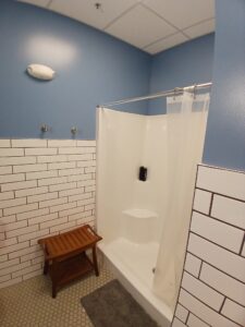 A bathroom with blue walls, white subway tile, hexagonal floor tiles, a white shower with a curtain, a wall-mounted soap dispenser, a stool, and a gray bath mat.
