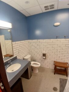 A bathroom with blue walls, white subway tile, a sink with a blue countertop, a toilet, a wooden bench, hooks on the wall, and hexagonal floor tiles. A mirror and light are above the sink.