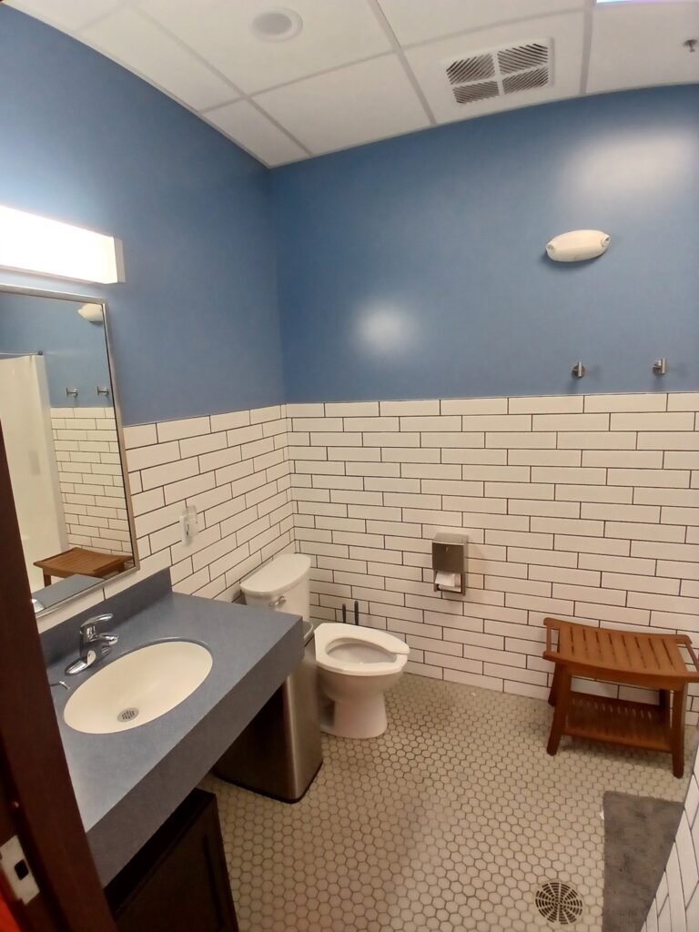 A bathroom with blue walls, white subway tile, a sink with a blue countertop, a toilet, a wooden bench, hooks on the wall, and hexagonal floor tiles. A mirror and light are above the sink.