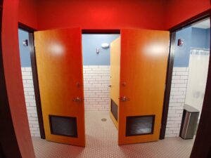 Two wooden restroom doors, both partially open, reveal tiled walls with white subway tiles, blue paint above the tiles, and hexagonal floor tiles. A shower curtain and trash can are visible on the right.