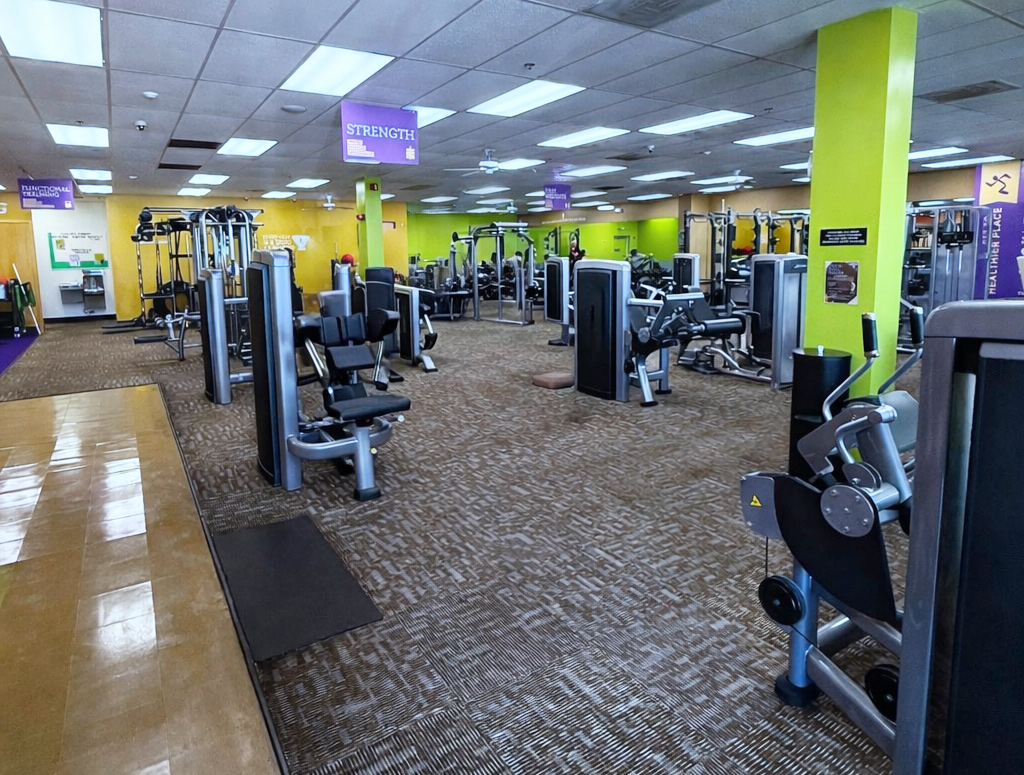 A spacious, brightly lit gym with yellow and green walls, featuring various weight machines and exercise equipment neatly arranged on a patterned carpet. Purple signs hang from the ceiling, labeling workout areas.