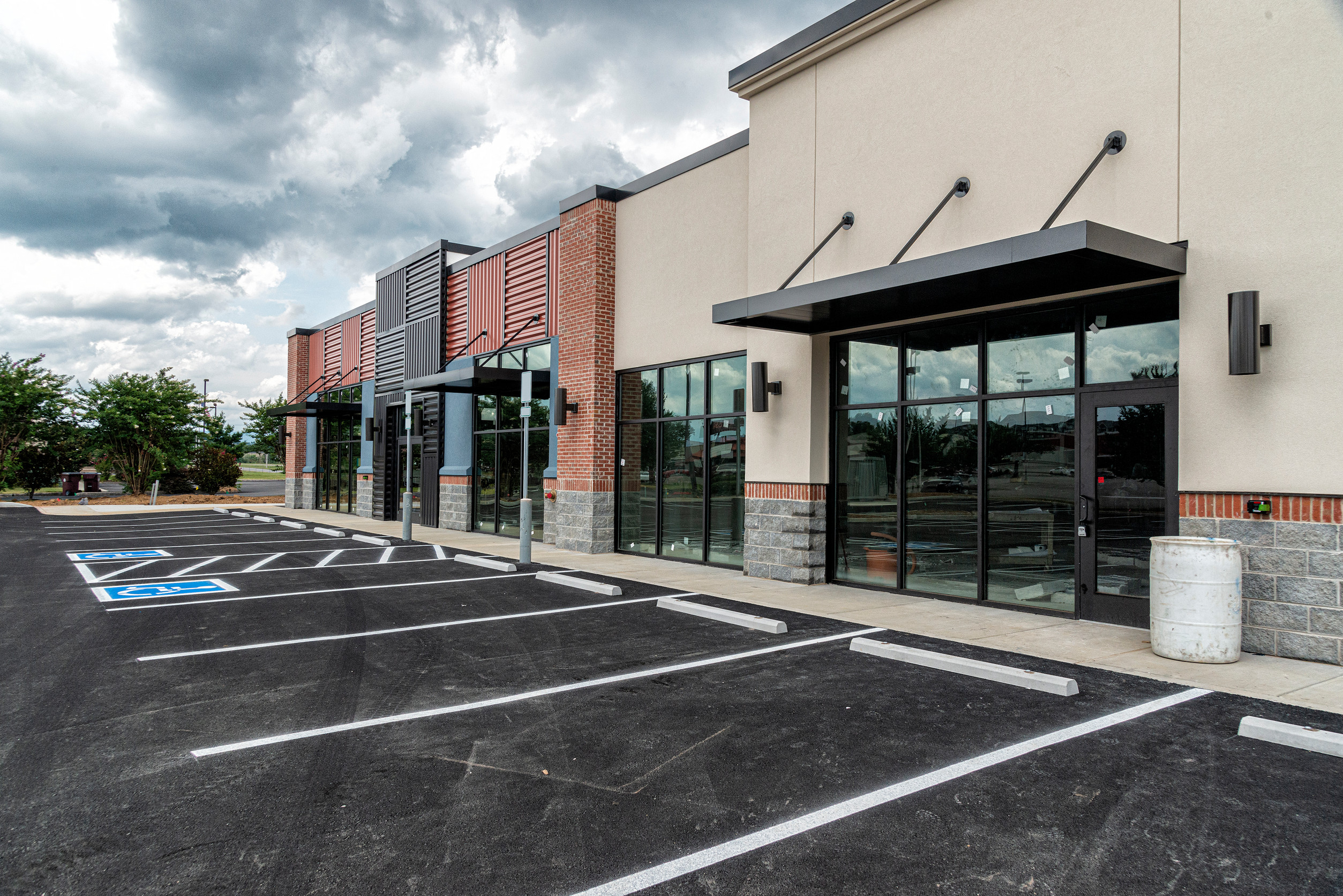 A newly constructed, empty strip mall with large glass storefronts, brick and beige exterior, and an empty parking lot featuring several accessible parking spaces under a cloudy sky.