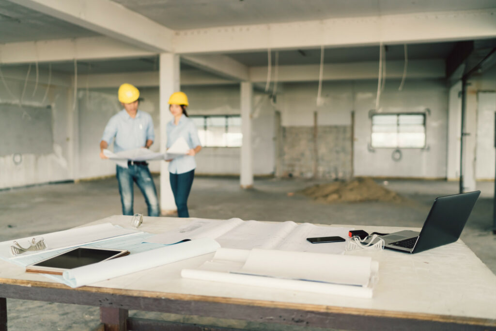 A table with blueprints, a tablet, and a laptop in the foreground; in the background, two people wearing yellow hard hats review plans inside an unfinished building.