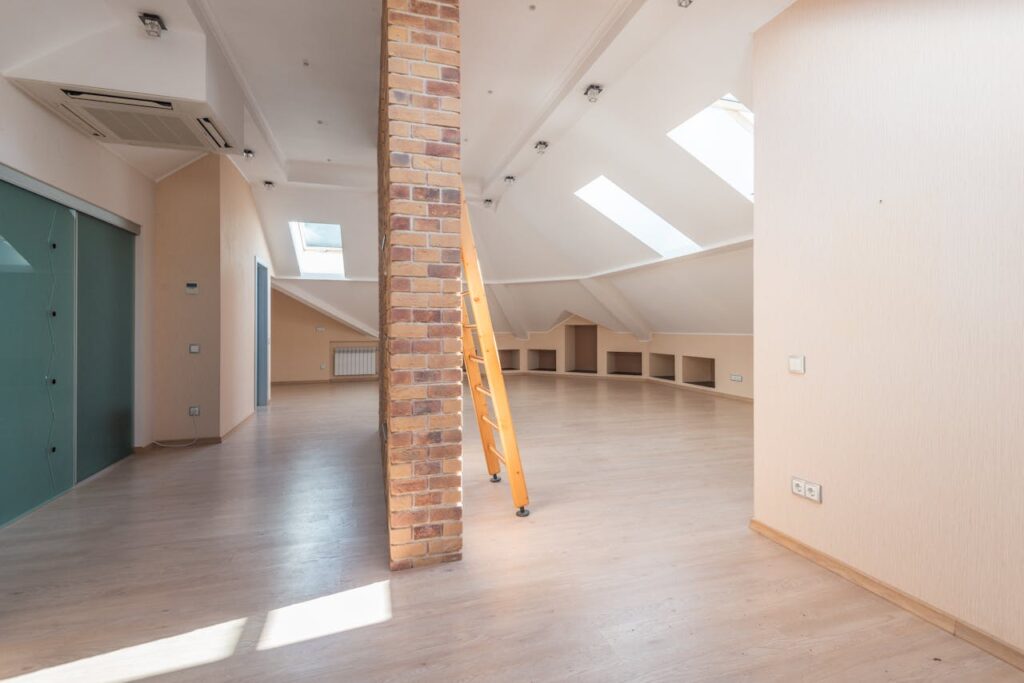 A spacious, modern attic room with light wood floors, beige walls, a brick column in the center, skylights, a yellow ladder, and built-in shelving under sloped ceilings.